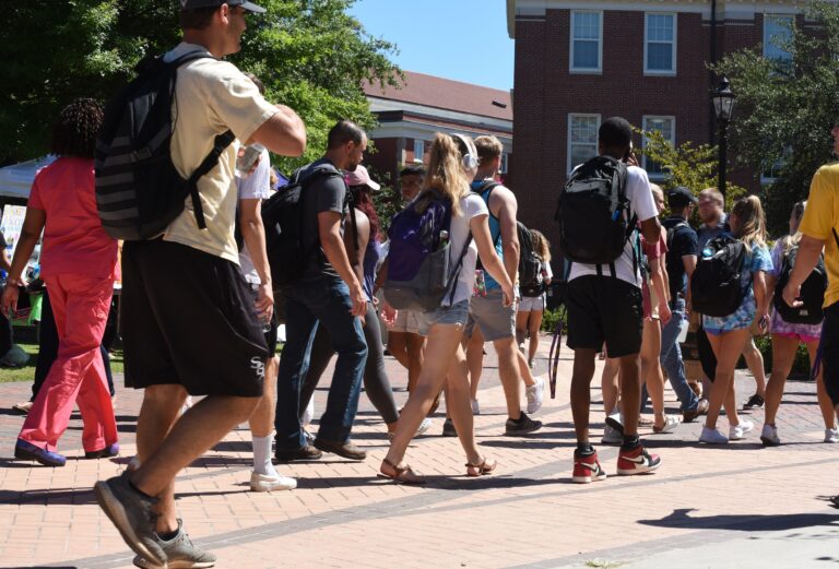 UNCC Finals Tips: The Ultimate Guide for 49ers Students 2 A diverse group of college students with backpacks walks across a brick paved walkway on a sunny campus day, with brick dormitories in the background.