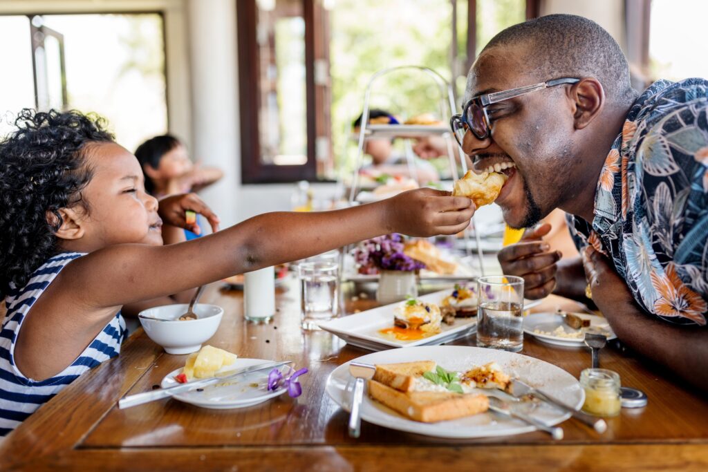 A young girl playfully feeds a piece of pastry to her smiling father during a bright family brunch at a restaurant table filled with breakfast dishes.